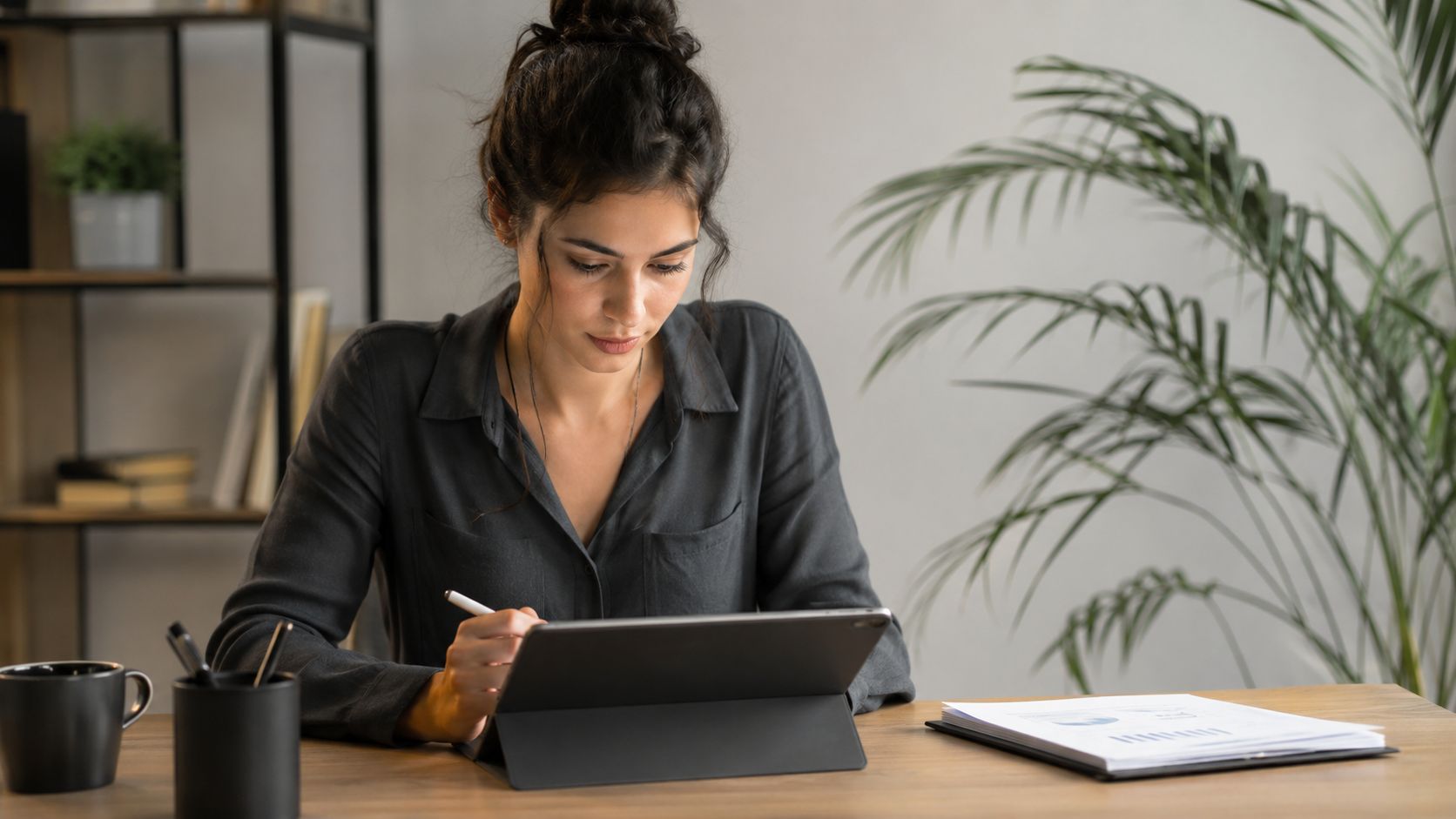 Una mujer joven con cabello recogido trabajando con una tableta y un lápiz óptico en su escritorio.